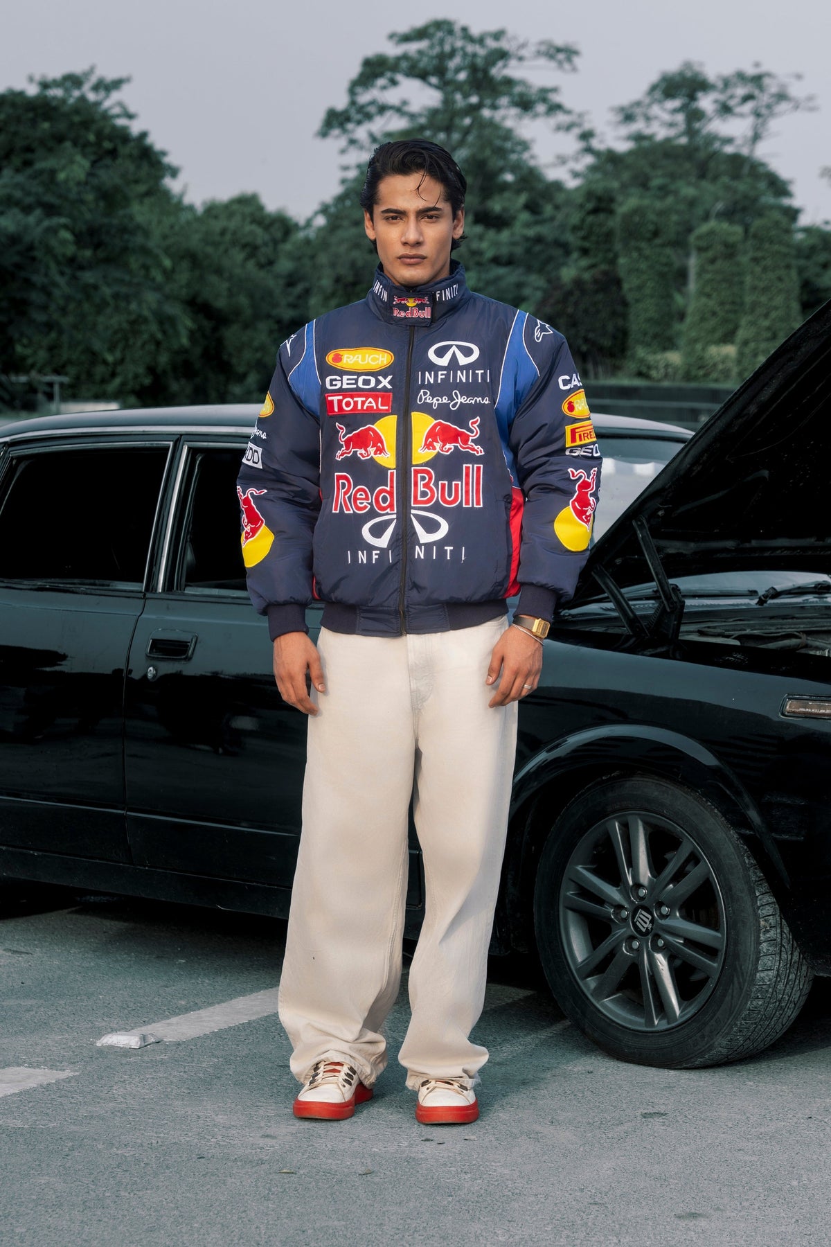 Koti Apparels Red Bull racing jacket with embroidered sponsor logos, worn by a man standing next to a black car.