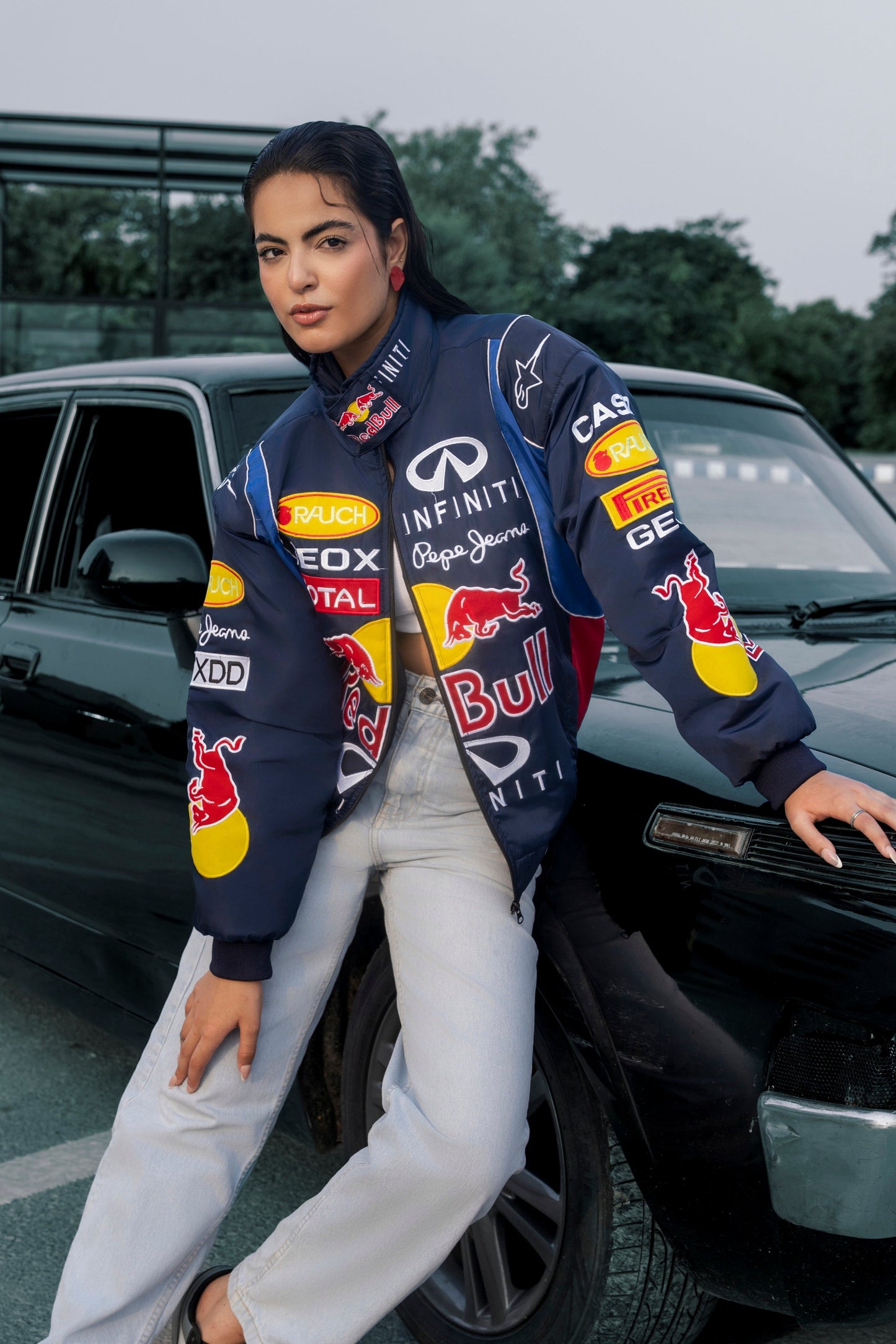 Model wearing a Koti Apparels navy blue Red Bull racing jacket with embroidered sponsor logos, leaning on a car.
