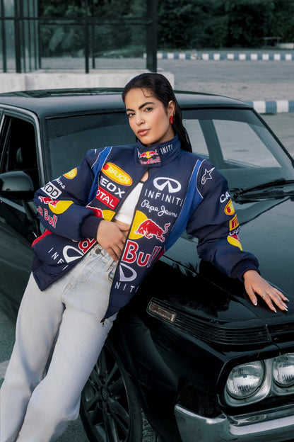 Model wearing a Koti Apparels navy blue Red Bull racing jacket with embroidered sponsor logos, leaning on a car.