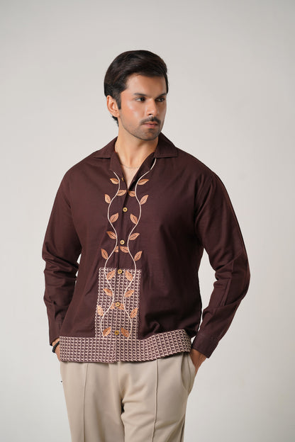 Man wearing a brown embroidered shirt with beige pants on a plain background