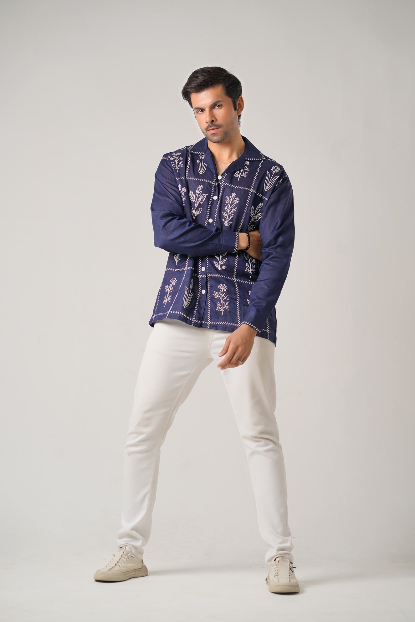 Man wearing a dark blue embroidered shirt and white pants on a gray background