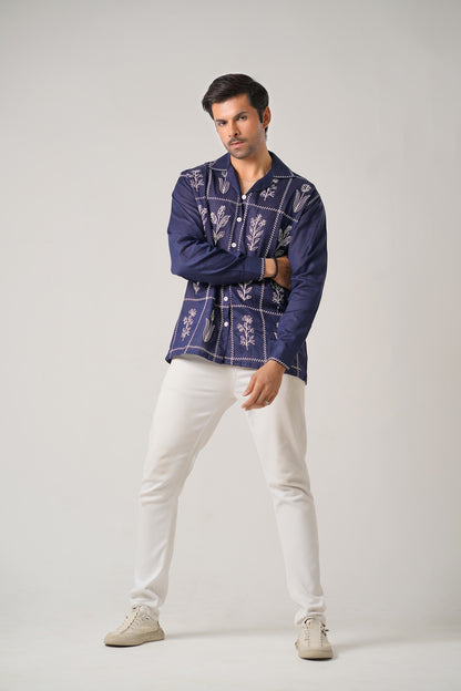 Man wearing a dark blue embroidered shirt and white pants on a gray background