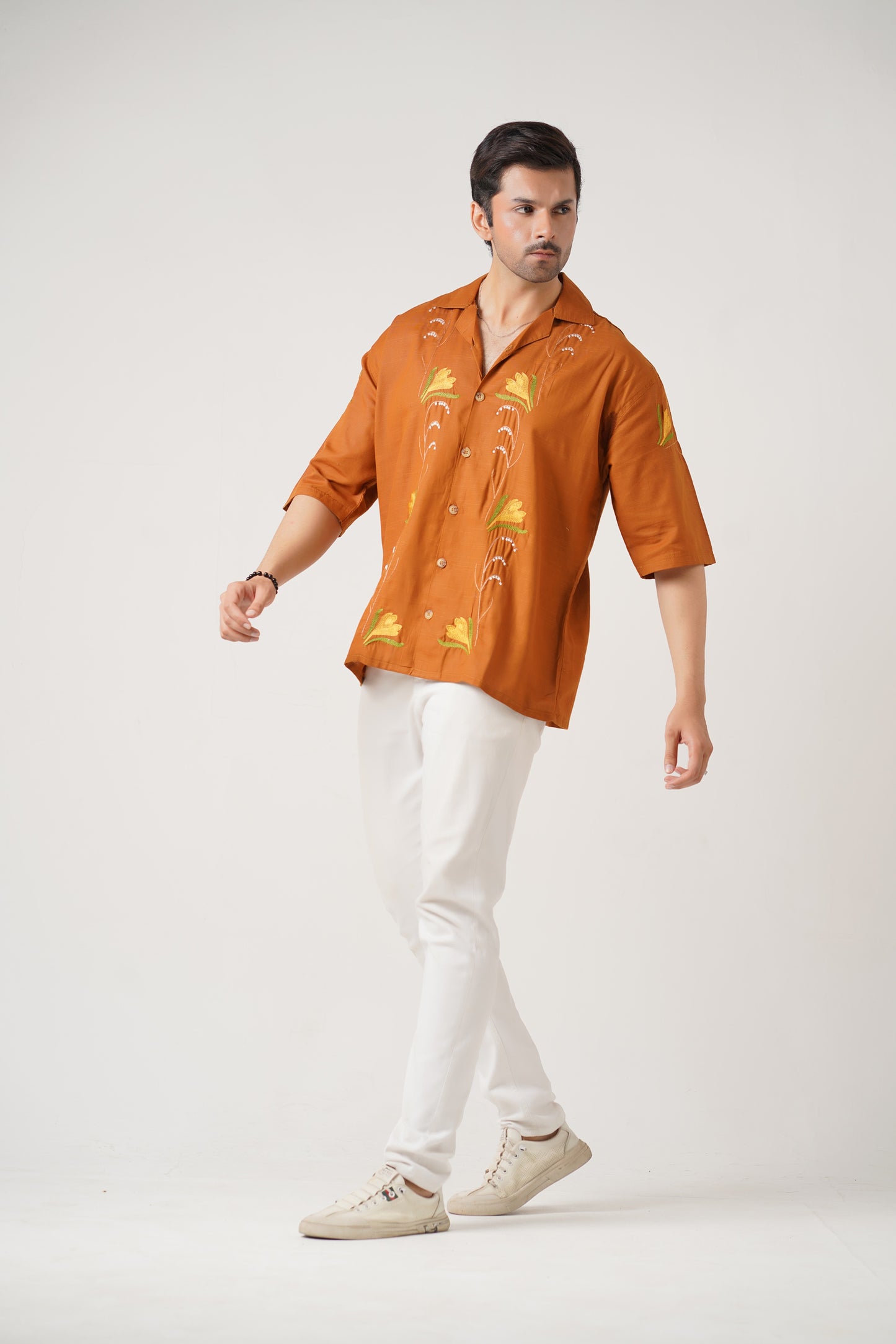 Man wearing a rust-colored shirt with gold patterns and white pants on a light gray background