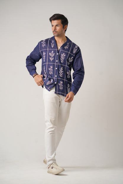 Man wearing a blue patterned shirt and white pants on a plain background