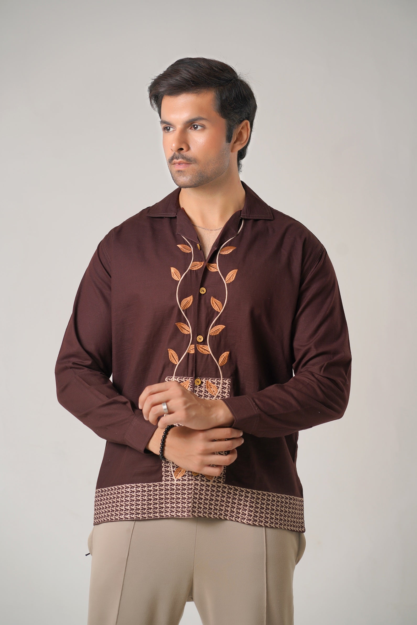Man wearing a brown embroidered shirt with beige pants on a plain background
